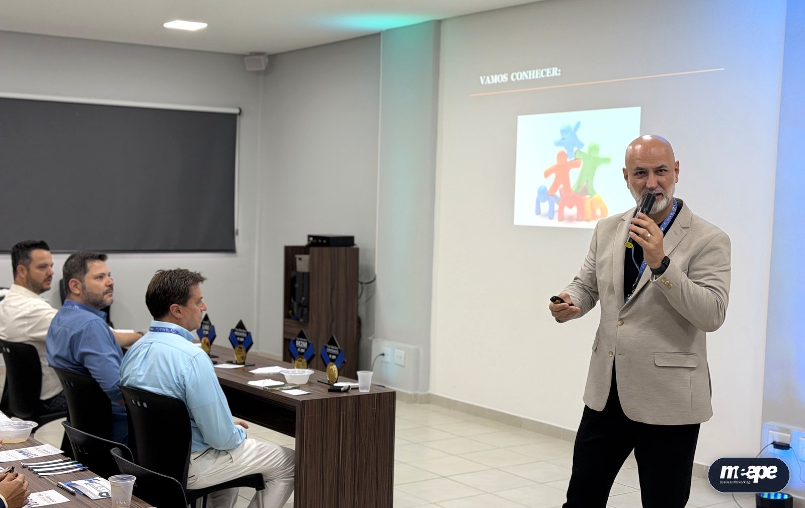 Donizete Claro falando ao microfone diante dos membros do Meepe, com o slide Vamos conectar ao fundo